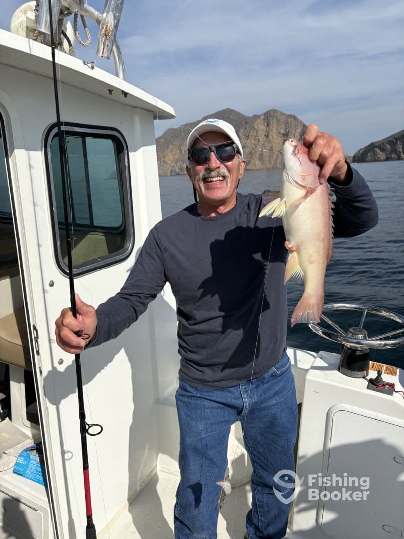 An enthusiastic angler proudly displaying a colorful fish aboard a fishing boat, with scenic rocky cliffs in the background.