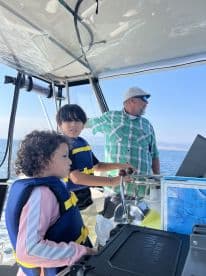 Two children in life jackets exploring the helm of a fishing boat, with an adult guiding them on a sunny day at sea.