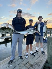 A family proudly displaying their catch of the day, featuring a large Redfish and a Bluefish, at a scenic dock during sunset.