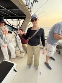 A woman proudly displaying a large Speckled Trout while fishing on a charter boat, with fellow anglers in the background.