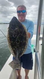 Angler proudly displaying a large Flounder while on a fishing trip, with a scenic water backdrop.