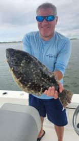 Angler proudly displaying a large Flounder while fishing on a boat, showcasing a successful day on the water.