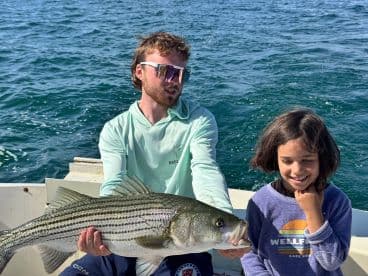 A young angler and a child proudly display a large Striped Bass while fishing on a boat, showcasing a successful day on the water.