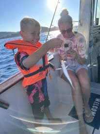 A young boy in a life jacket proudly holds a small fish while fishing with an adult on a boat, enjoying a sunny day on the water.
