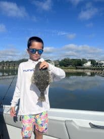 A young angler proudly displaying a Flounder while fishing from a boat in a scenic coastal environment.
