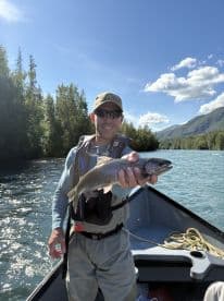 A proud angler holding a Rainbow Trout while fishing from a boat in a scenic river setting.