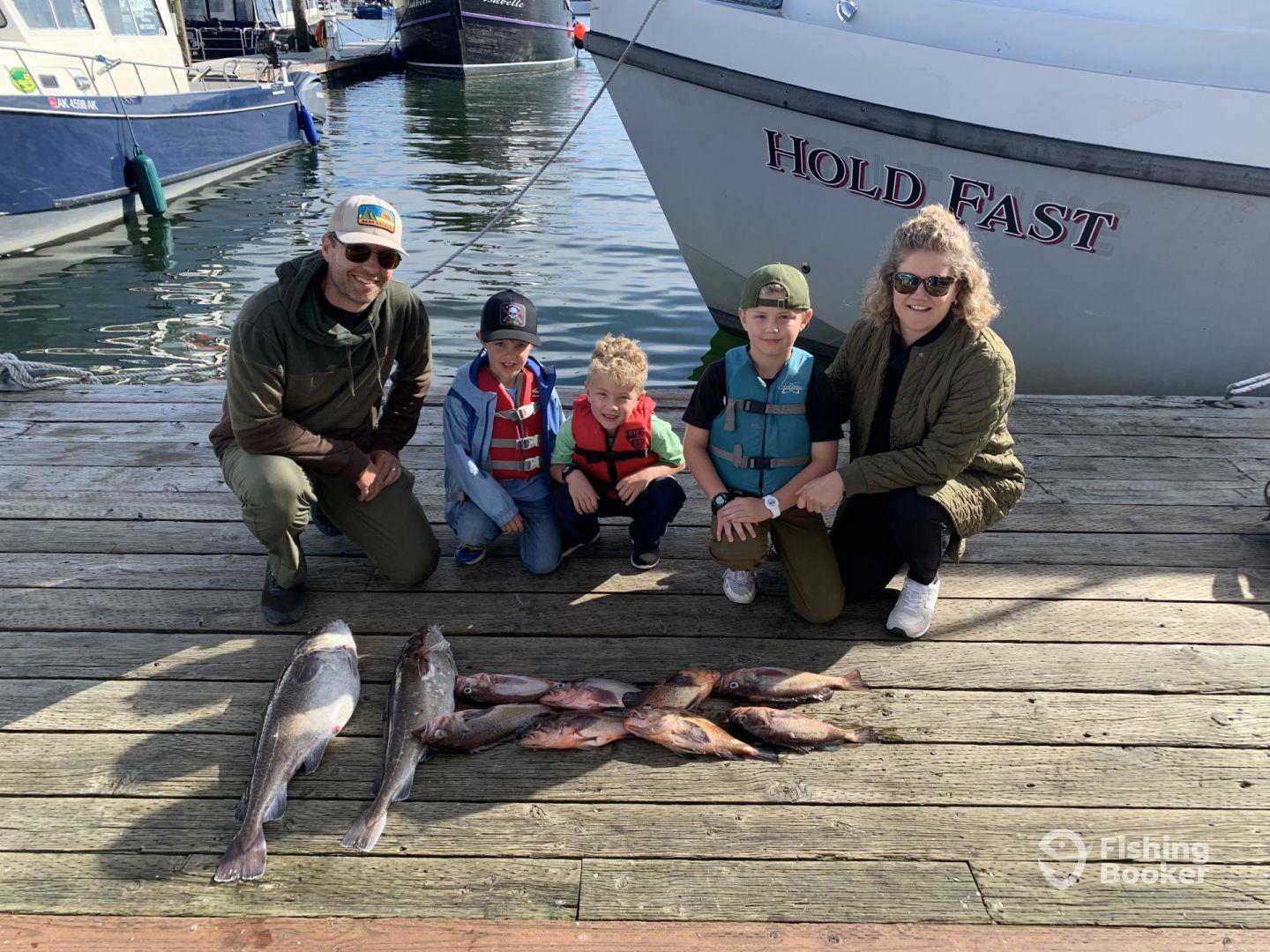 A family proudly displaying their catch of the day, featuring several fish, at the dock next to the boat named 'Hold Fast'.