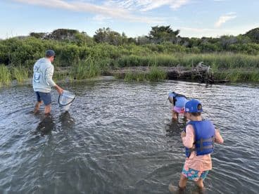 A family enjoying a shallow water fishing experience, with adults and children exploring the area while wearing life jackets.
