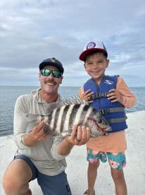 A young boy proudly holds a striped Sheepshead fish alongside an adult angler at the dock, showcasing a successful fishing trip.