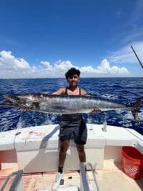 A young angler proudly displaying a large Wahoo aboard a fishing boat in clear blue waters.