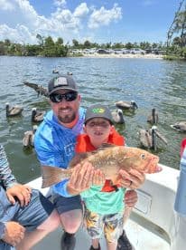 A young boy and his father proudly displaying a large Grouper while fishing in a scenic coastal environment, surrounded by pelicans.