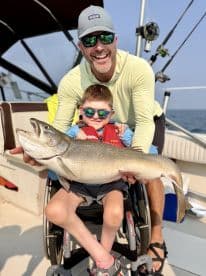 A young boy in a wheelchair proudly holds a large Lake Trout with the help of an adult on a fishing boat.