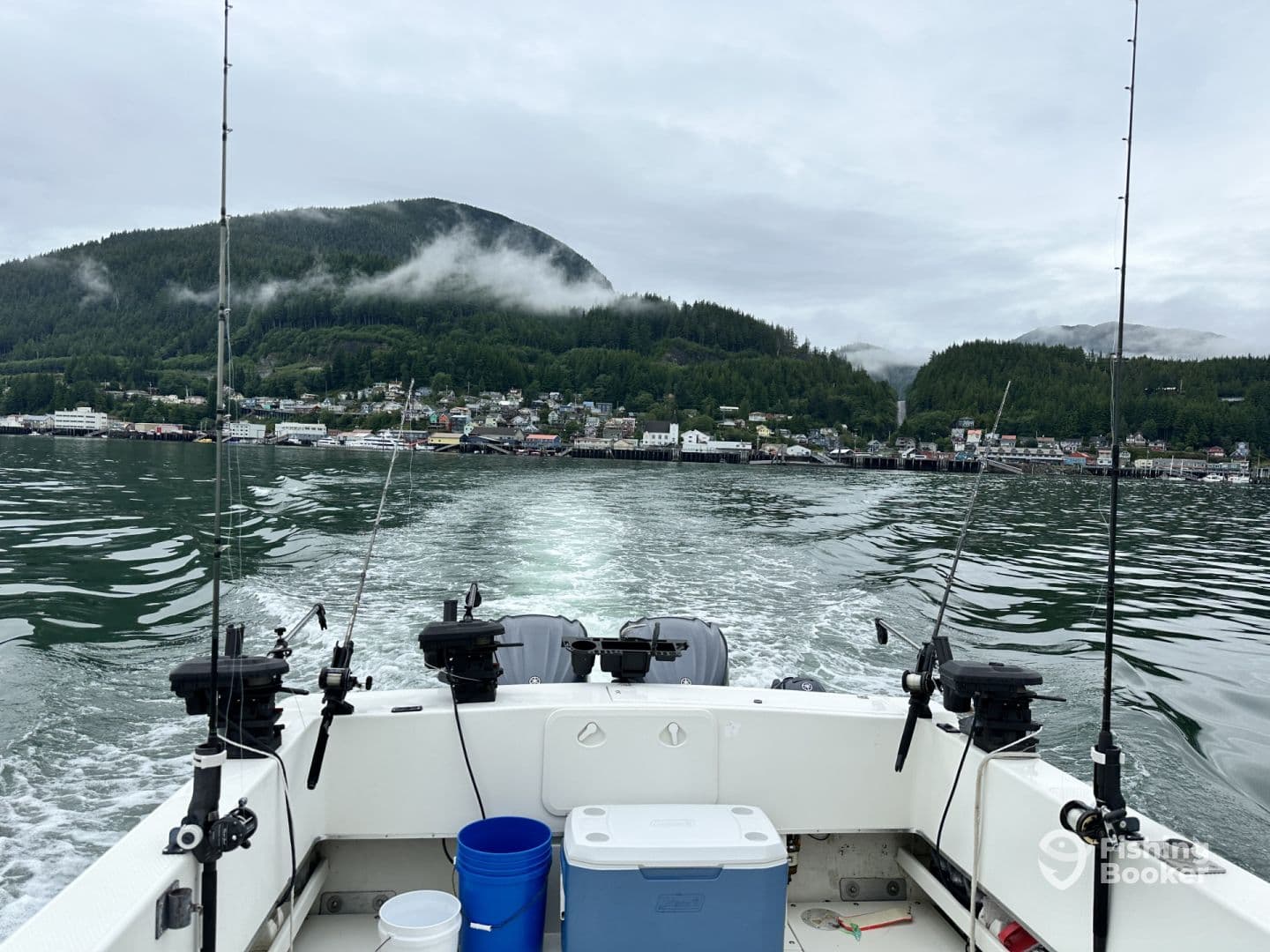 View from the interior of a fishing boat cruising through a scenic coastal area, showcasing fishing rods and a cooler against a backdrop of lush mountains and a small town.