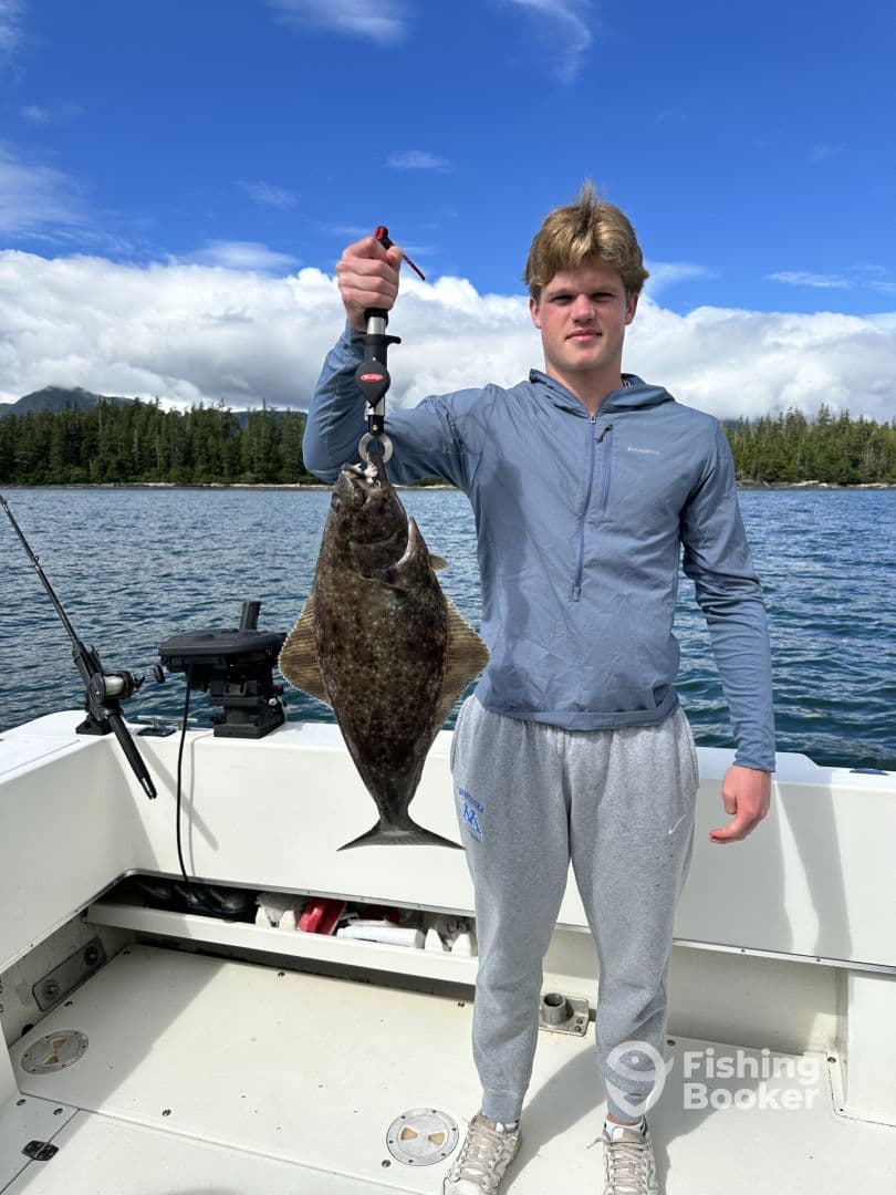 A young angler proudly displaying a large Halibut while fishing on a boat in a scenic coastal environment.