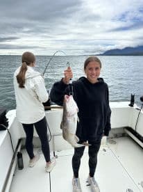 Young angler proudly holding a large Cod while fishing on a boat, with another person actively reeling in a catch in the background.