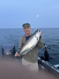 An angler proudly displaying a large Salmon while fishing on a boat in open water, with a serene evening sky in the background.