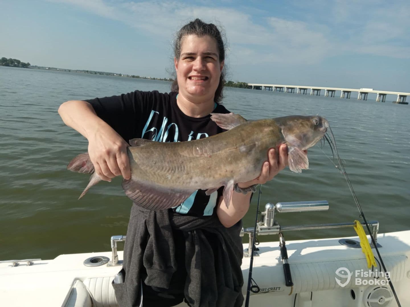 An angler proudly displaying a large Catfish while fishing on a boat, with a scenic water backdrop.