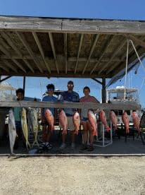 A family proudly displays their catch of the day, featuring multiple Red Snapper and a Mahi Mahi, at a fishing dock.