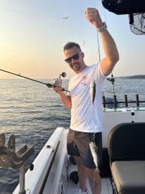 Angler reeling in two small fish while fishing on a boat during sunset.