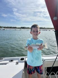 A young angler proudly displaying a Speckled Trout while fishing from a boat in a coastal environment.