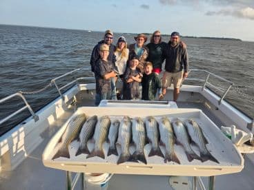 A family group poses on a fishing boat, showcasing a successful catch of striped bass arranged neatly on a board.
