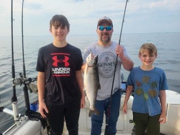 A family fishing trip on a boat, showcasing a proud moment with a caught fish, likely a trout, in a serene water setting.