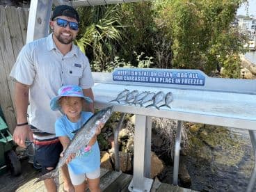 A young girl proudly displays a large King Mackerel while standing next to an adult at a fish cleaning station, showcasing their successful catch from a recent fishing trip.
