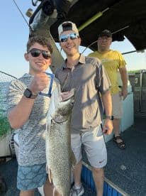 Two anglers proudly displaying a large Lake Trout on a fishing charter, with a third person in the background. The setting appears to be on a boat, showcasing a successful fishing trip.
