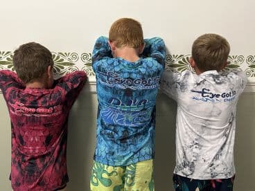 Three young boys wearing colorful fishing shirts from 'Eye Got Ya Charters' pose with their backs to the camera, showcasing their attire.