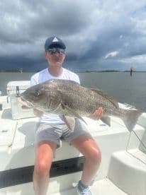 A young angler proudly displaying a large Black Drum while fishing on a boat, with a stormy sky in the background.