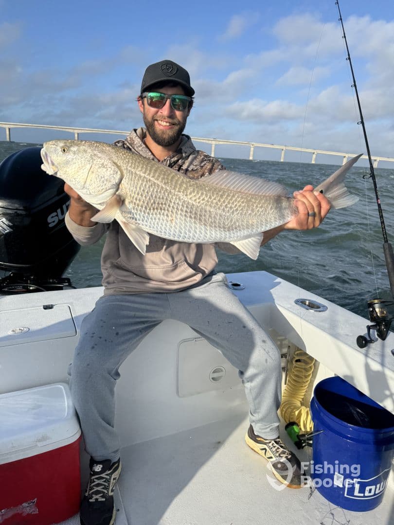 An angler proudly displaying a large Redfish while fishing from a boat, with a scenic bridge in the background.