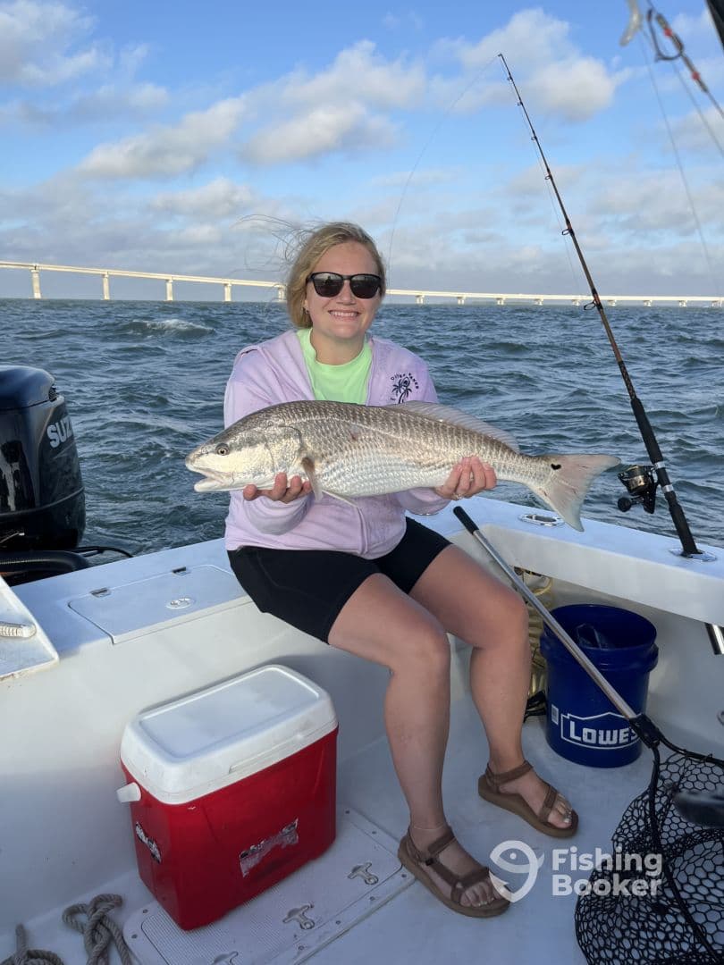 An angler proudly displaying a large Redfish while fishing on a boat, with a scenic bridge in the background.