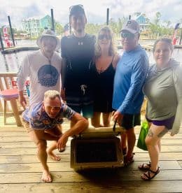 A group of five friends gathered around a cooler on a dock, showcasing their catch after a fishing trip.
