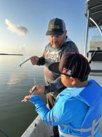 A young boy in a life jacket watches intently as an adult demonstrates how to handle a small fish while fishing on a calm morning.
