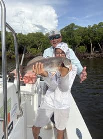 A young angler proudly displaying a large Redfish while fishing from a boat in a mangrove setting.