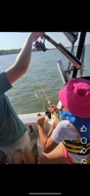 A young girl in a life jacket is actively reeling in a fish while an adult assists her on a boat, showcasing a fun fishing experience.