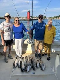 A family group proudly displaying their catch of multiple Lake Trout on a fishing charter, with a scenic lighthouse in the background.