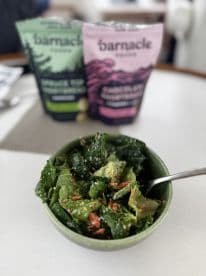 A bowl of salad with two bags of Barnacle food products in the background, set on a dining table.
