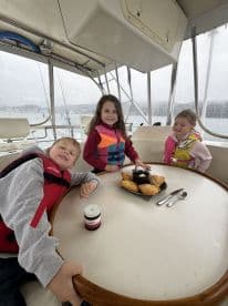 Three children enjoying snacks inside the cabin of a boat, showcasing a cozy atmosphere on a rainy day.