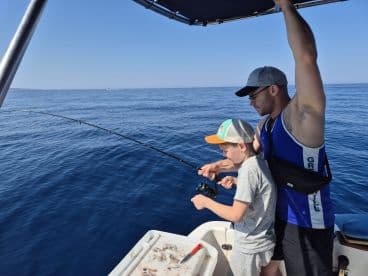 A young boy and an adult are engaged in fishing on a boat, with the boy reeling in a line while the adult assists, showcasing a bonding moment on the open water.