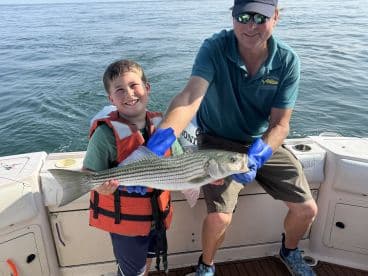 A young angler and an adult proudly displaying a large Striped Bass on a fishing boat, showcasing a successful day on the water.
