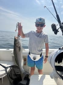 Young angler proudly holding a large Striped Bass while fishing on a boat, showcasing a successful day on the water.