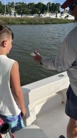 A young boy watches intently as an adult shows him a freshly caught fish while fishing from a boat in a scenic marina.