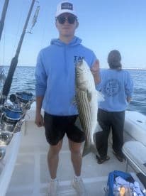 A young angler proudly displaying a striped bass while fishing on a boat, with another person in the background.