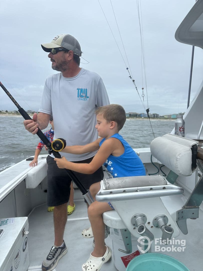 A young boy is actively reeling in a fishing line while being guided by an adult on a fishing boat, showcasing a family fishing experience.