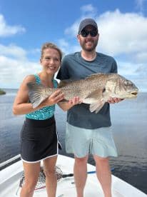 Two anglers proudly displaying a large Black Drum while fishing on a boat in a scenic waterway.