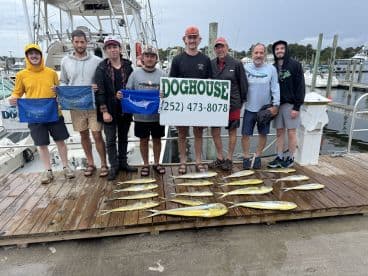 A group of anglers proudly displaying their catch of Mahi Mahi on a dock, with the Doghouse charter boat in the background.