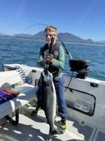 A young angler proudly displaying a large Salmon while fishing on a boat in a scenic coastal environment.