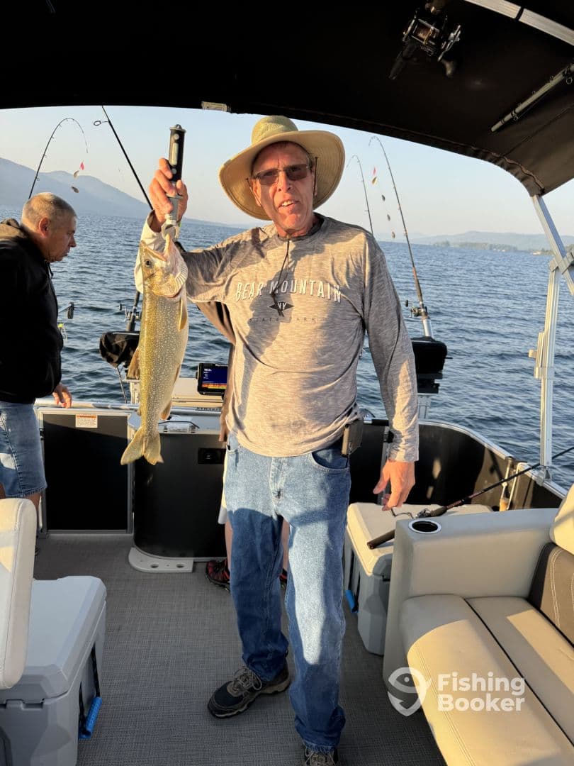 An angler proudly displaying a large Lake Trout while fishing on a boat, surrounded by scenic water and mountains.