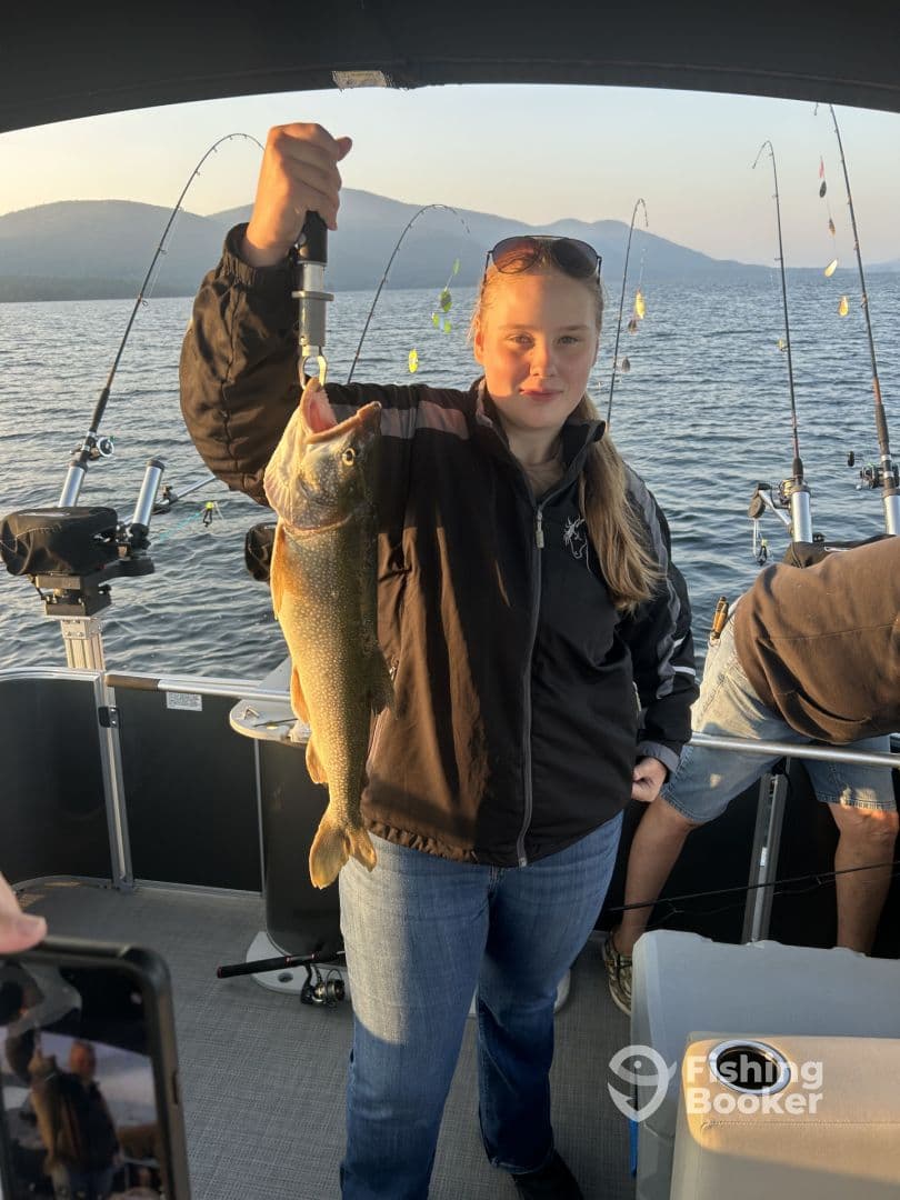 A young angler proudly displaying a large Lake Trout while fishing on a scenic lake, surrounded by fishing rods and a beautiful mountainous backdrop.